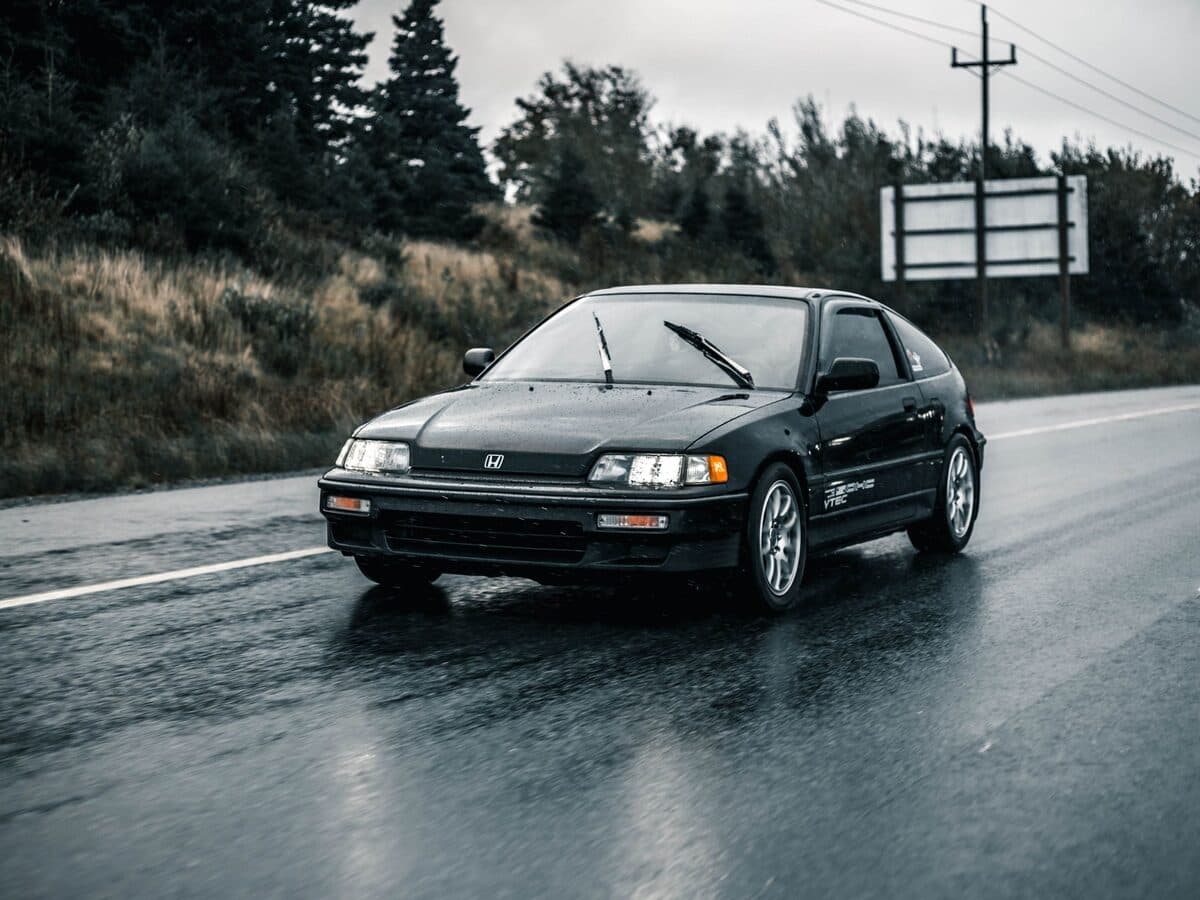 A black Japanese hatchback on a wet road lined with trees